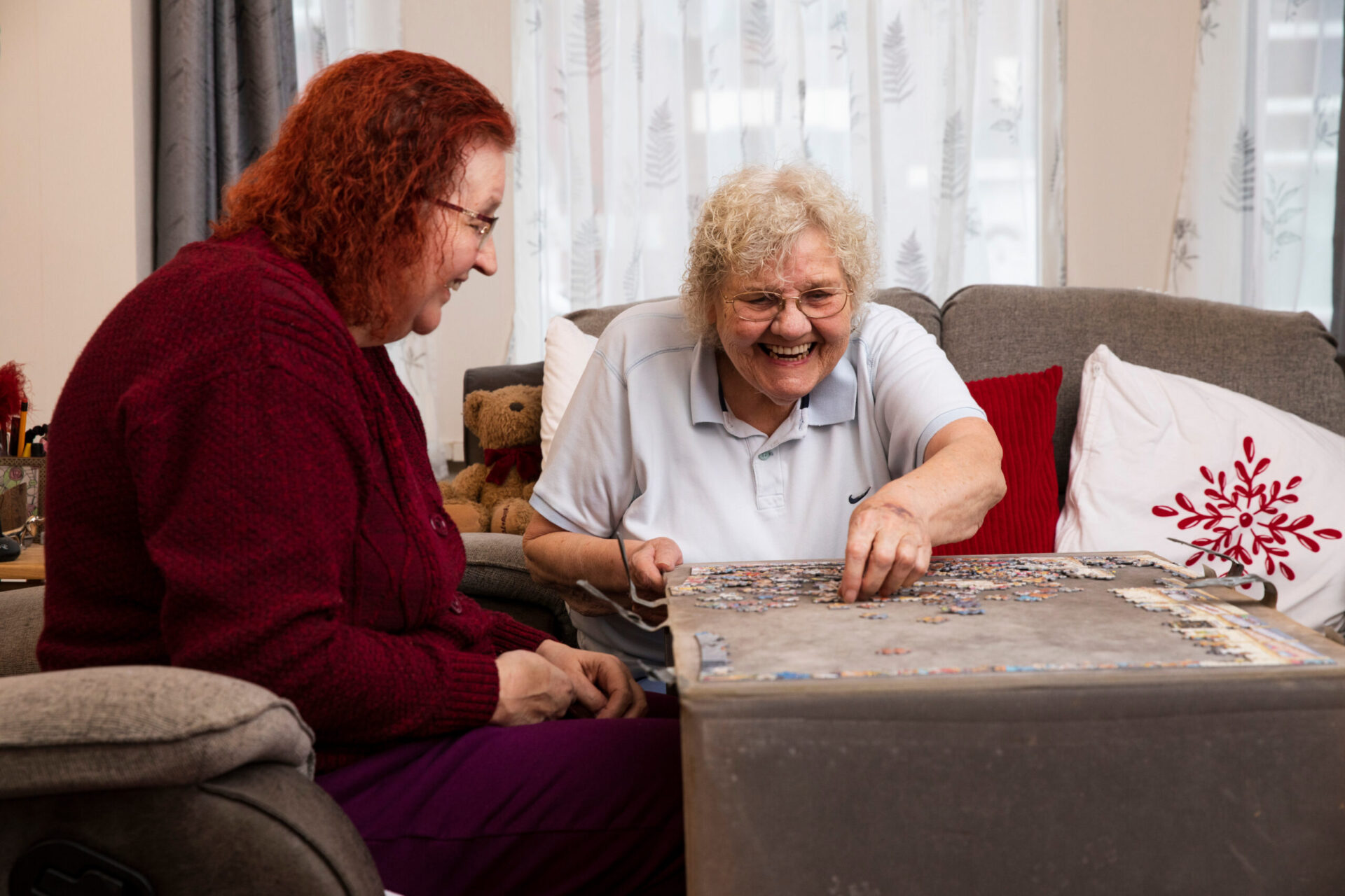 Two older people are smiling as they sit on a sofa completing a jigsaw puzzle together.