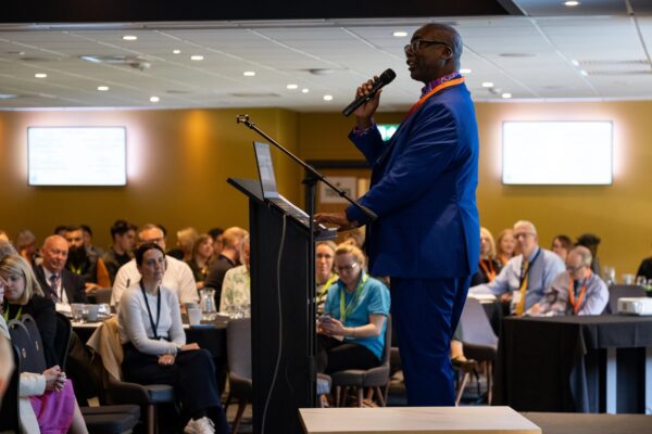 Patrick Vernon on stage holding a microphone as he addresses the audience of the conference